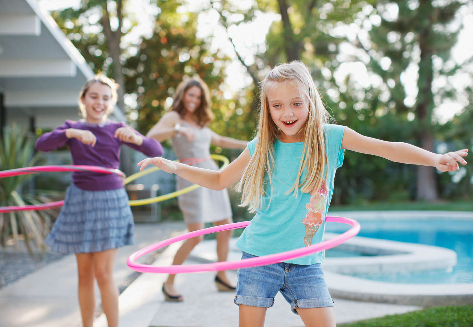 Young girl hula-hooping with a couple friends in a back yard by a pool