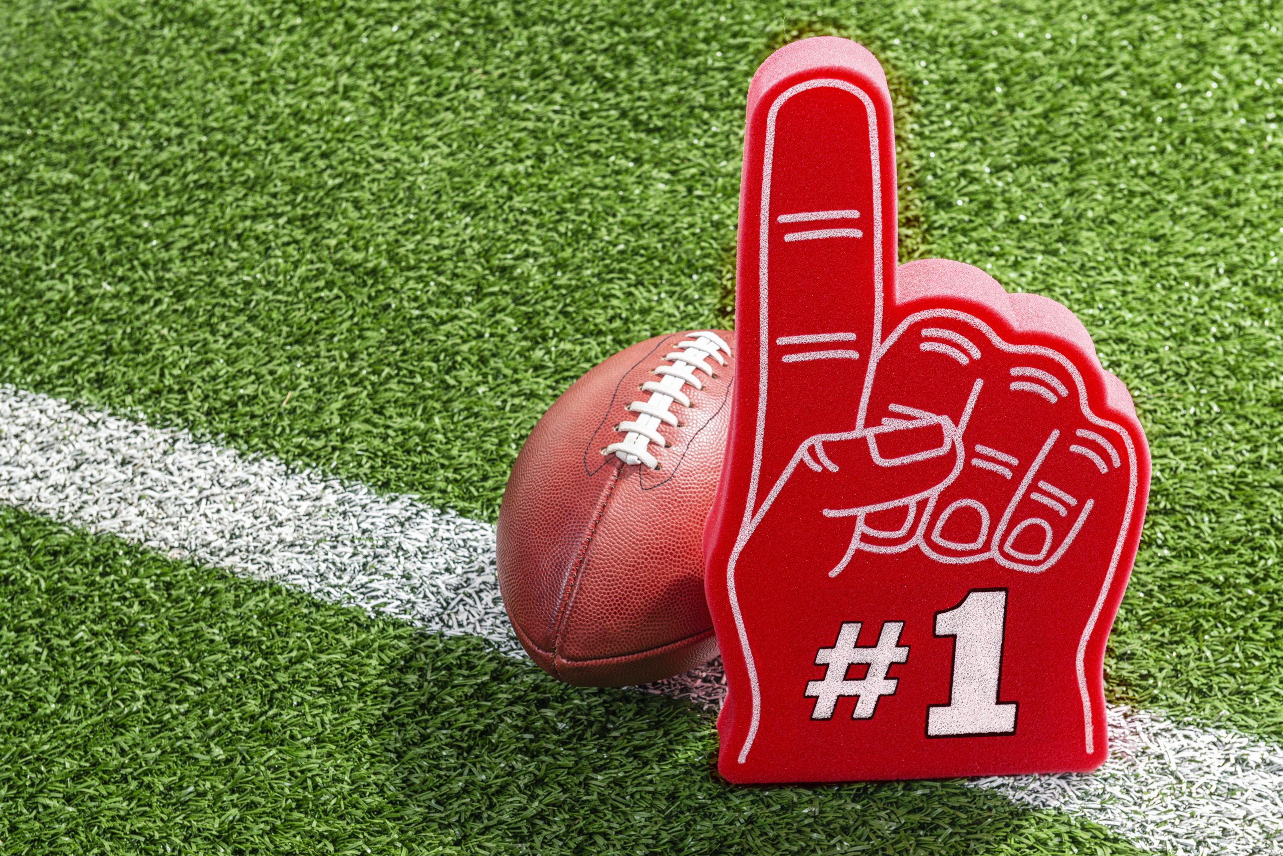 A red foam hand with a white #1 outlined in black sitting next to a leather American football that are both sitting on a white yard line on the stadium grass